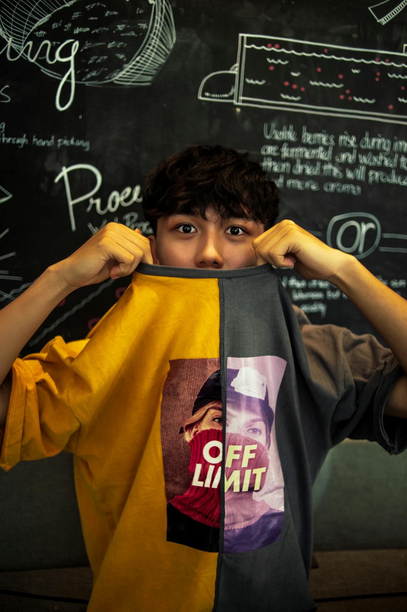 Asian teenager playfully posing with graphic shirt in front of chalkboard in classroom setting.