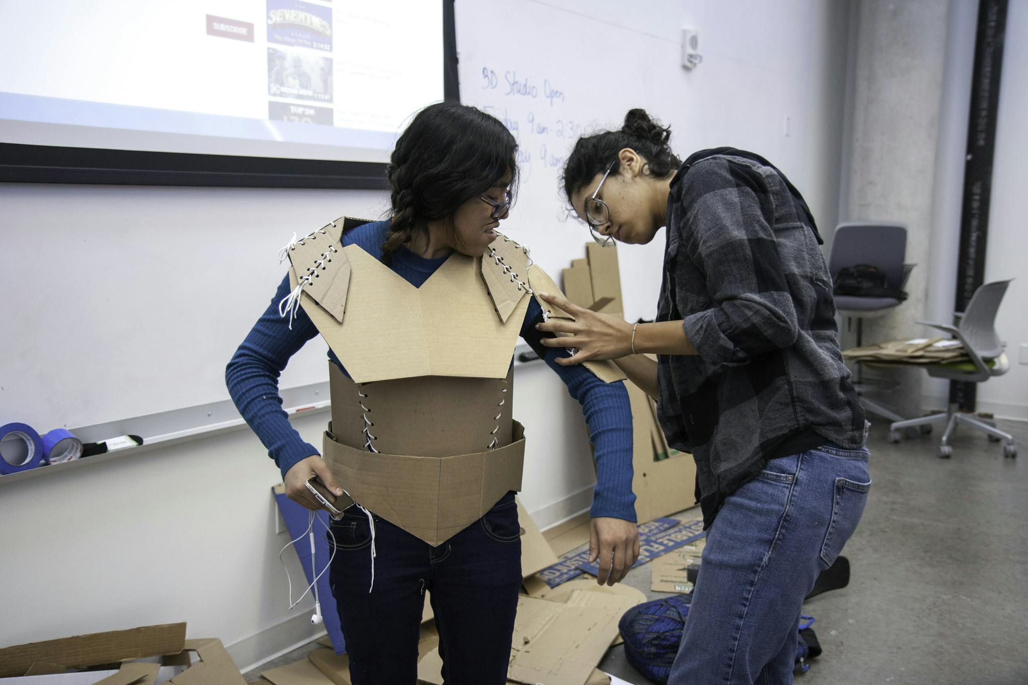 Two students collaborate on creating cardboard armor during a college art class project indoors.