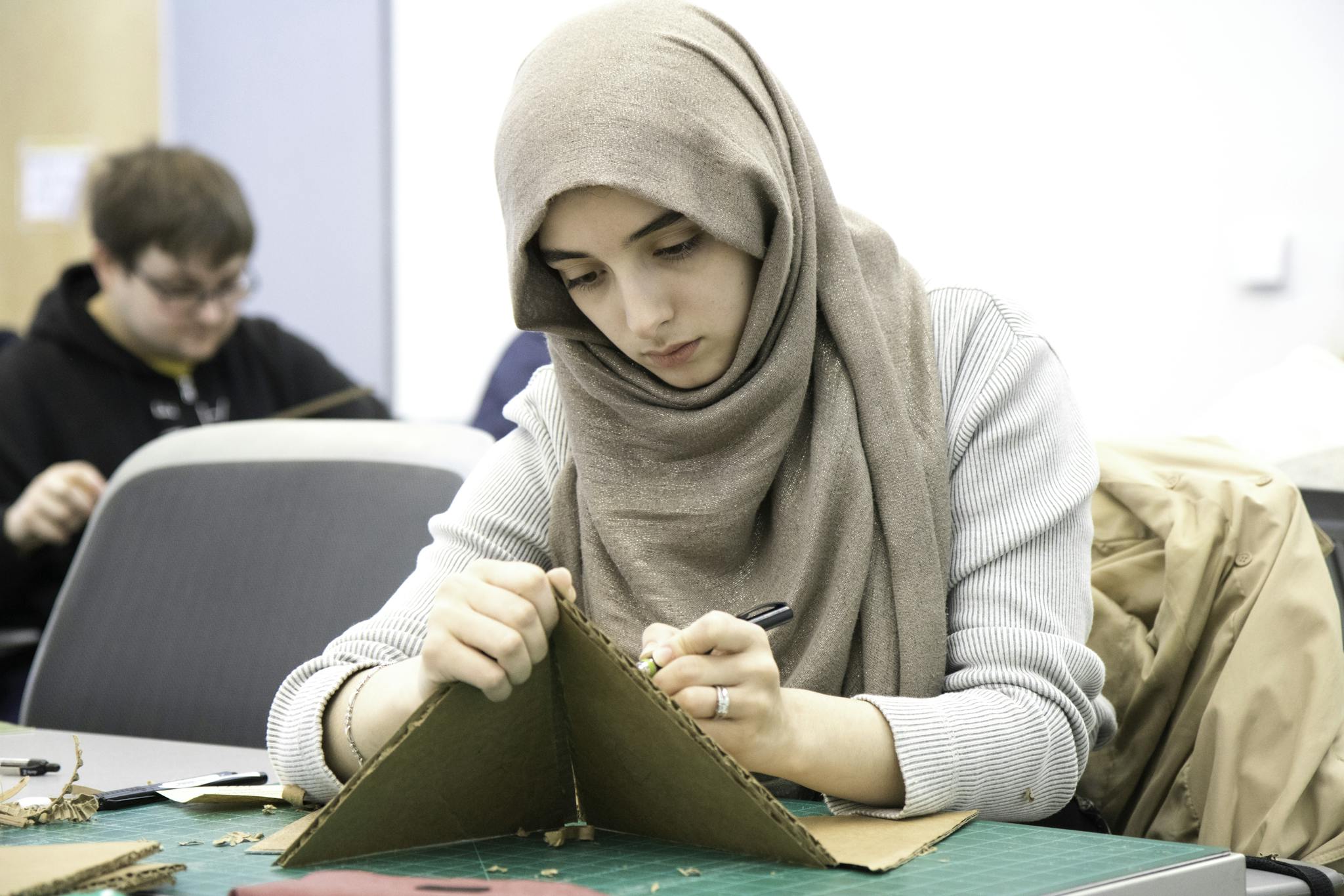 Young Muslim woman wearing hijab working on a cardboard craft project in classroom setting.